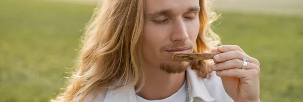 Jeune homme aux cheveux longs et les yeux fermés sentant palo santo parfumé bâton à l'extérieur, bannière — Photo de stock