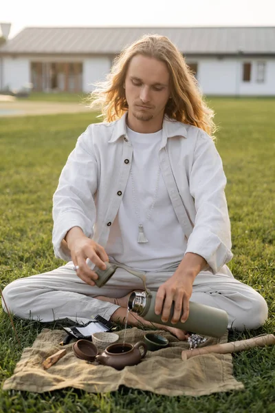 Long haired man in linen clothes pouring hot water from thermos into ceramic teapot — Stock Photo
