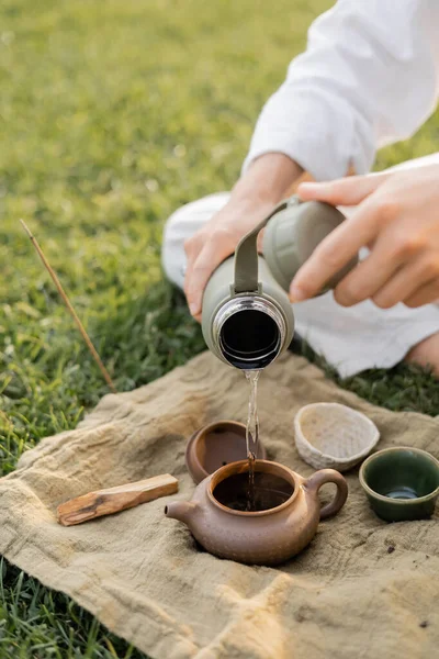 Abgeschnittene Ansicht von Yoga-Mann, der heißes Wasser in Keramik-Teekanne gießt, während er auf Rasen in der Nähe von Leinenteppich mit Tassen und Palo Santo Stick sitzt — Stockfoto