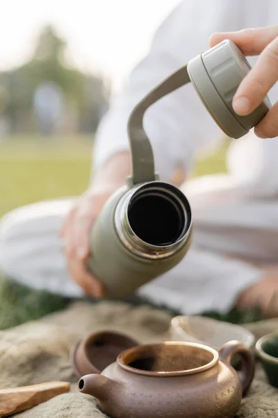 Частичный вид размытого человека, держащего термос возле глиняного чайника во время чайной церемонии на открытом воздухе — стоковое фото