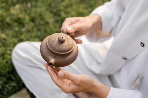 Cropped view of man in white linen clothes holding clay teapot while sitting outdoors — Stock Photo