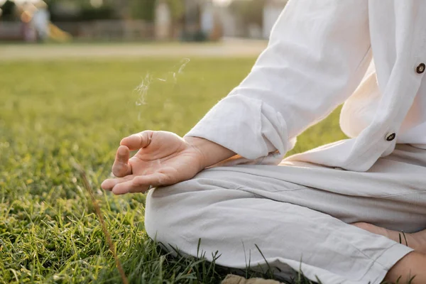 Частичный вид человека в белой одежде, сидящего в удобной позе йоги и медитирующего возле ароматного дыма на травянистой лужайке — стоковое фото