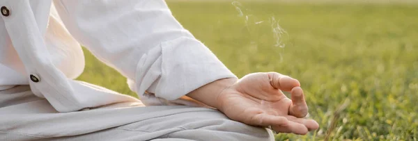 Vista recortada del hombre en ropa de lino que muestra gyan mudra gesto mientras medita cerca de humo perfumado en el césped verde, pancarta - foto de stock