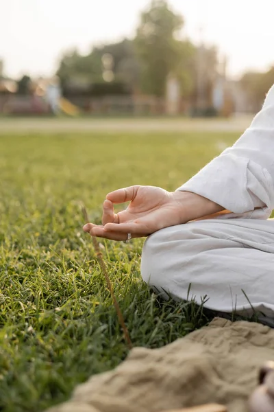 Vista recortada del hombre en ropa de lino blanco meditando con gesto de gyan mudra mientras está sentado en el campo de hierba verde - foto de stock