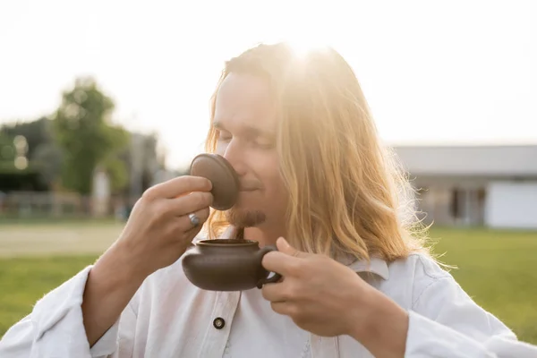 Jovem com cabelos longos e olhos fechados desfrutando de aroma de chá puer branco segurando bule oriental ao ar livre — Fotografia de Stock