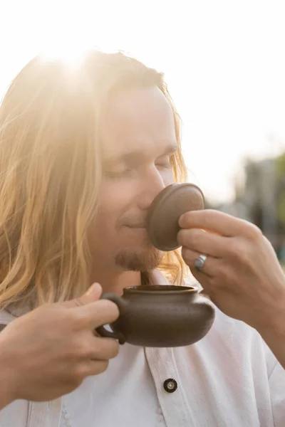Pleased long haired man with closed eyes holding oriental teapot and enjoying flavor of puer tea in sunshine outdoors — Stock Photo