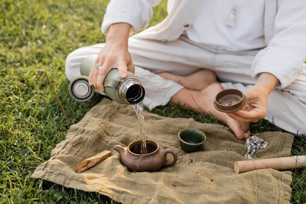 Teilansicht von Yoga-Mann, der heißes Wasser in Lehm-Teekanne gießt, während er auf dem Rasen in der Nähe von Leinenteppich mit Mala-Perlen und Palo Santo-Stick sitzt — Stockfoto