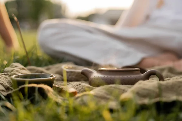 Linen rug with ceramic teapot and cups on green grass near man in blurred background — Stock Photo