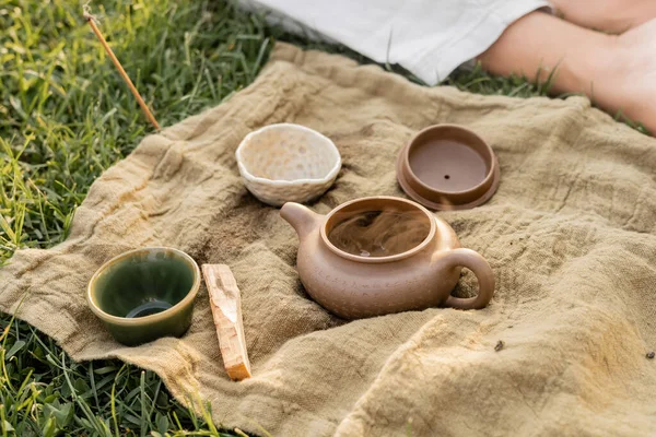 Aromatic palo santo stick and ceramic teapot with cups near cropped yoga man sitting on grassy lawn — Stock Photo