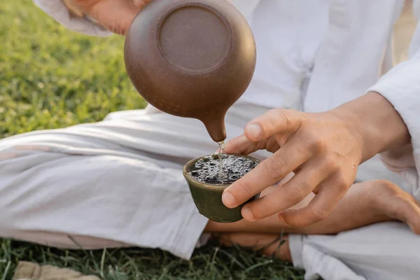 Vista parcial do homem de ioga em roupas de linho sentado no gramado verde e derramando chá puer de bule oriental em tigela de barro — Fotografia de Stock