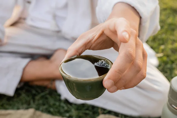 Vue rapprochée de tasse d'argile avec du thé puer dans la main de l'homme de yoga cultivé assis à l'extérieur — Photo de stock