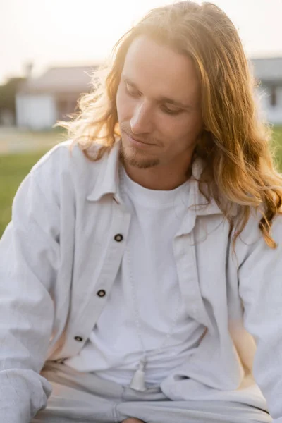 Joven y soñador hombre con el pelo largo y claro meditando mientras está sentado al aire libre - foto de stock