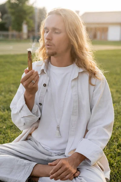 Длинноволосый мужчина в белой льняной одежде, дующий в ароматный пало-санто во время эзотерического ритуала на открытом воздухе — стоковое фото
