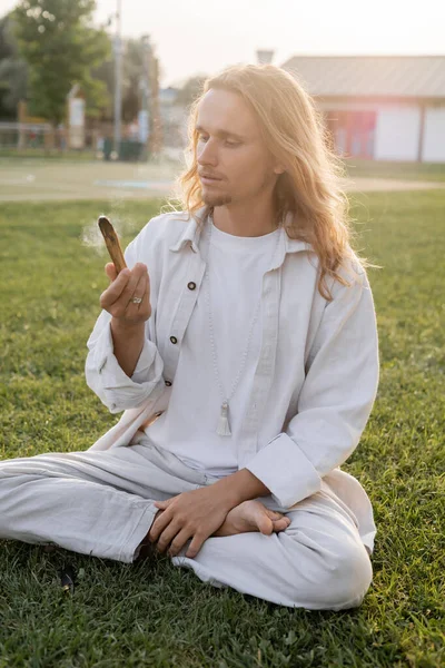 Стильный мужчина в белой льняной одежде сидит в легкой позе и держит тлеющий пало Санто придерживаться во время медитации на открытом воздухе — стоковое фото
