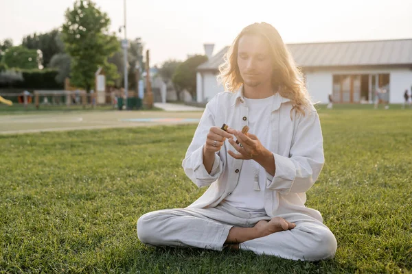 Long haired man in white linen clothes sitting in easy yoga pose and holding lighter and aromatic palo santo stick outdoors — Stock Photo