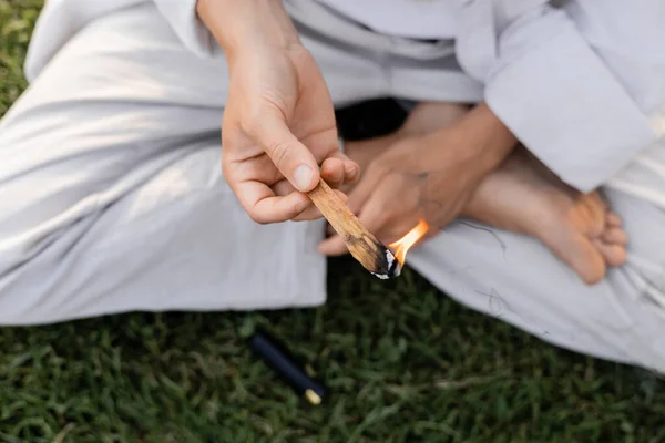 Ansicht von oben beschnittener Mann mit brennendem Palo Santo Stick, während er im Freien in lockerer Yoga-Pose sitzt — Stockfoto