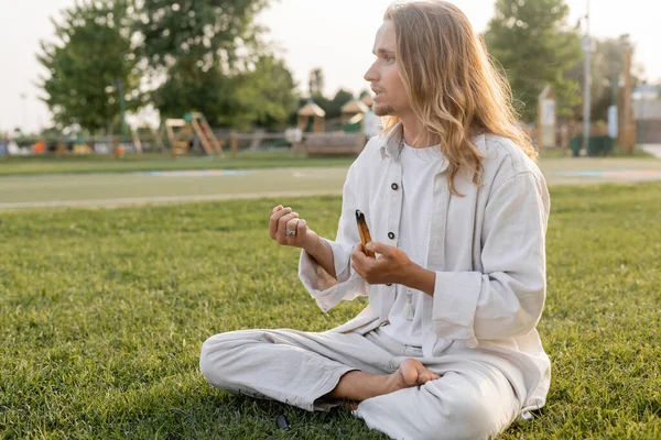Barefoot man in white linen clothes meditating in easy pose with smoldering palo santo stick and looking away outdoors — Stock Photo