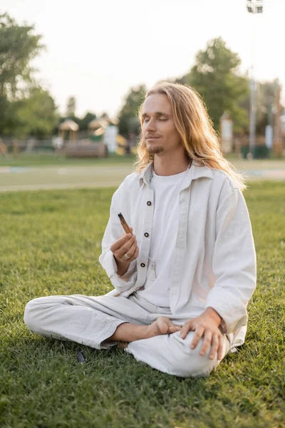 Hombre complacido en ropa blanca sosteniendo palo santo aromático palo durante la meditación en sukhasana pose sobre hierba verde - foto de stock