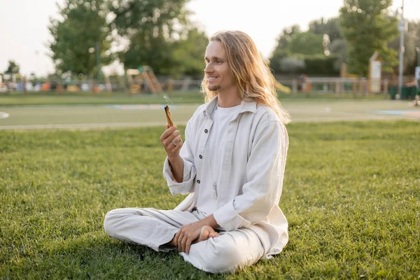 Счастливый длинноволосый мужчина с ароматным пало санто палкой во время медитации в легкой позе на зеленом газоне — стоковое фото
