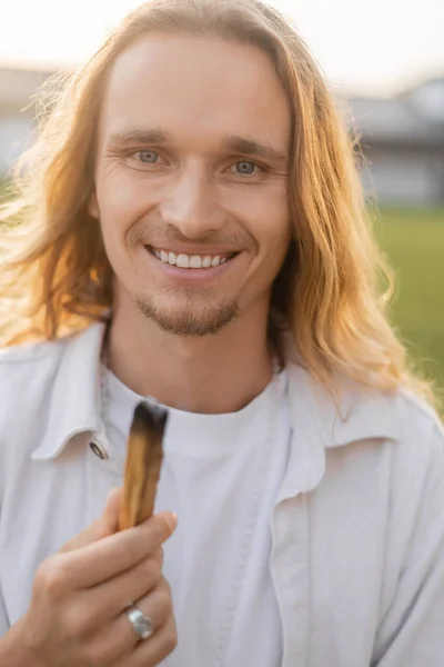 Радостный длинноволосый йога-человек держит тлеющий пало санто палкой и улыбается в камеру на открытом воздухе — стоковое фото