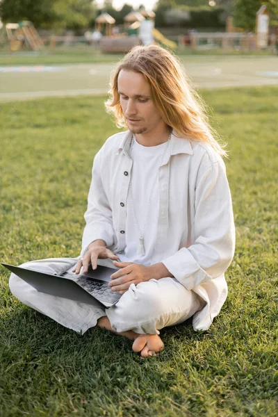Молодой длинноволосый мужчина в белой хлопчатобумажной одежде сидит в удобной позе и использует ноутбук на зеленой лужайке — стоковое фото