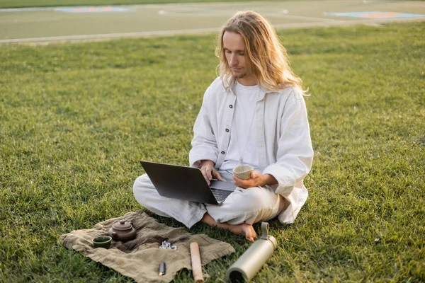 Молодий чоловік у білому одязі тримає чай Пуерто і використовує ноутбук, сидячи поруч з термосом і керамічним чайником на зеленому газоні — стокове фото