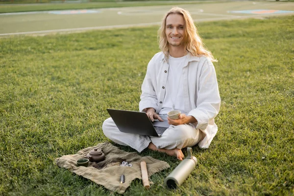 Homem de cabelos longos feliz sentado com laptop e copo de barro perto de garrafa térmica e tapete de linho com bule no gramado gramado — Fotografia de Stock