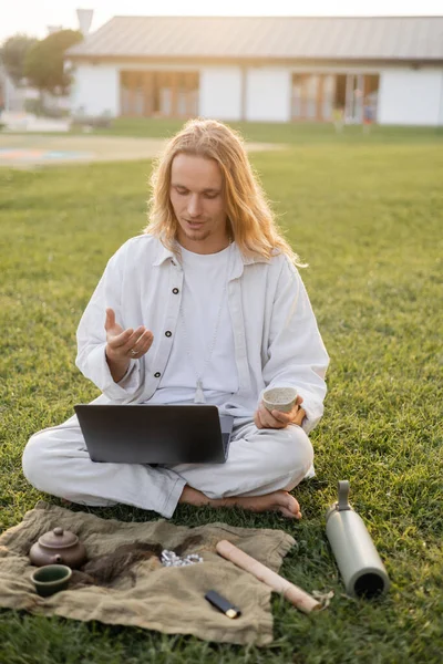 Hombre de pelo largo en ropa de lino que tiene videollamada en el ordenador portátil mientras está sentado con té puro cerca de la alfombra con taza de arcilla y tetera en el césped verde - foto de stock