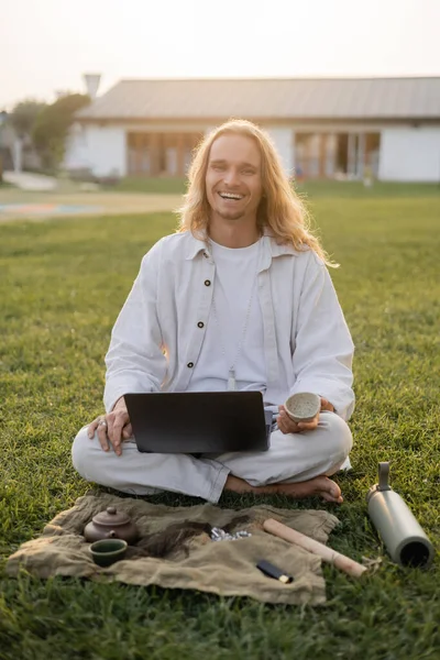 Carefree man smiling at camera while sitting on green grass with laptop and clay cup near thermos and linen rug with teapot — Stock Photo