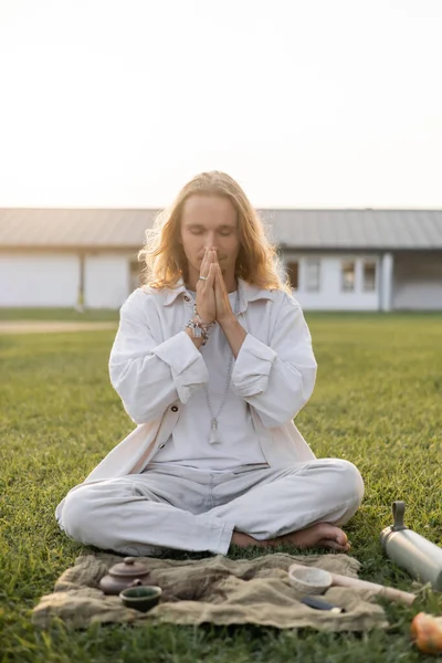 Young man in white clothes meditating in easy pose near linen rug with clay teapot and bowls on green lawn — Stock Photo