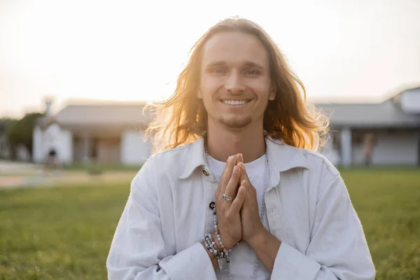 Carefree long haired man with mala beads showing anjali mudra gesture and smiling at camera outdoors — Stock Photo