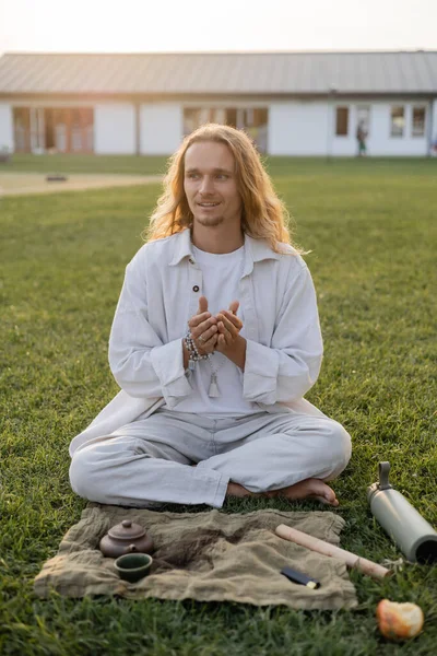 Homme aux cheveux longs souriants en vêtements blancs assis près du tapis de lin avec théière en céramique et regardant loin dans la campagne — Photo de stock