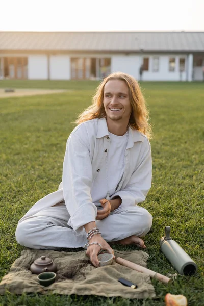 Heureux yoga homme en lin vêtements tenant tasse d'argile et regardant loin tout en étant assis sur la pelouse verte à l'extérieur — Photo de stock