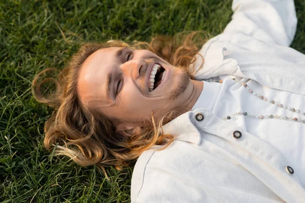 Vista ad alto angolo di felice uomo in camicia bianca e perline sdraiato su erba verde all'aperto — Foto stock