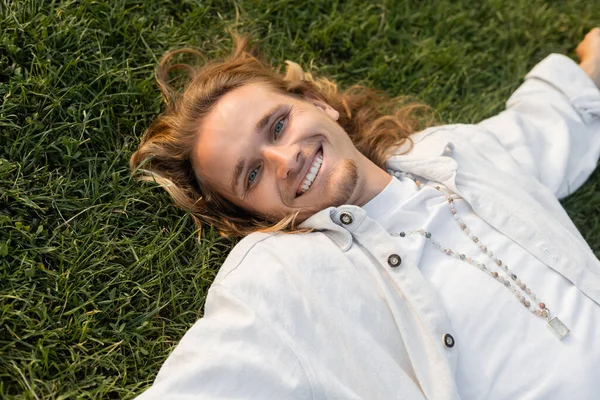 High angle view of cheerful young man in white linen shirt lying on green lawn and smiling at camera — Stock Photo
