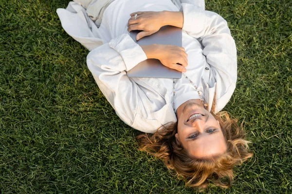 Top view of carefree man in white stylish clothes lying with laptop on green lawn — Stock Photo