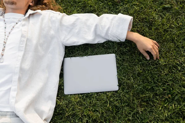 Vue du dessus de l'homme cultivé en chemise de coton blanc et des perles couchées sur l'herbe verte près de l'ordinateur portable — Photo de stock