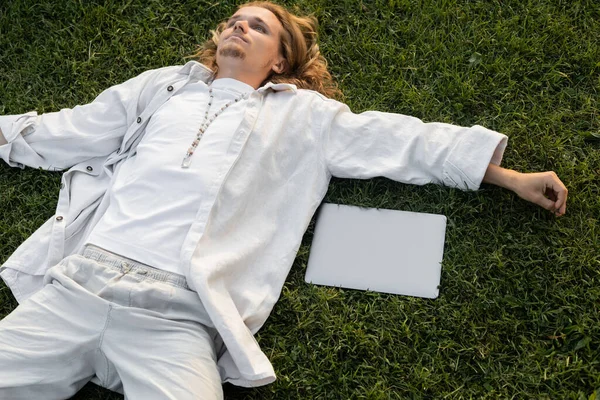 Vista de ángulo alto del hombre joven del yoga en ropa blanca que miente cerca del ordenador portátil en el campo verde - foto de stock