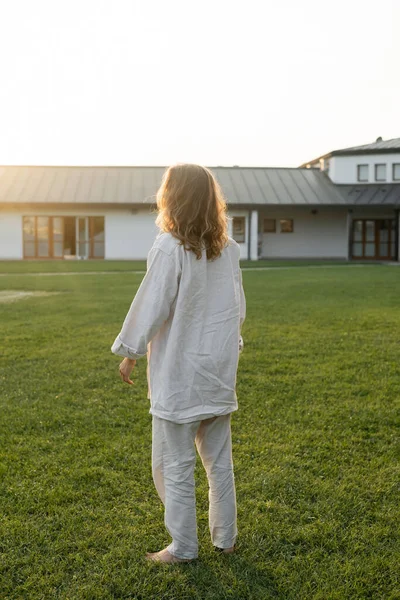 Back view of barefoot and long haired man in linen shirt and pants standing on green lawn near countryside house — Stock Photo