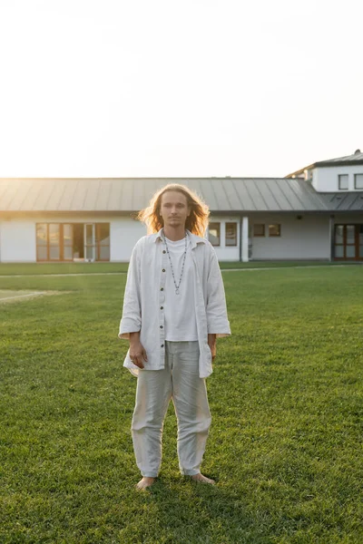 Full length of yoga man in cotton clothes standing on grassy lawn near cottage on background — Stock Photo