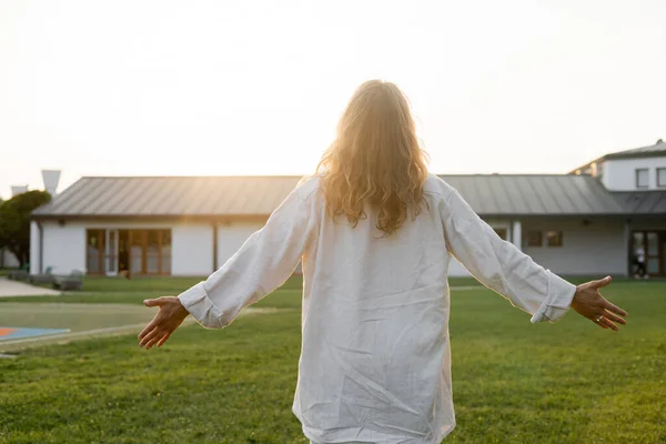 Back view of long haired man in white linen shirt meditating with outstretched hands near cottage outdoors — Stock Photo
