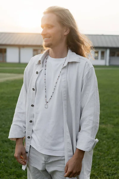 Joven hombre de yoga de pelo largo en camisa de lino y perlas sonriendo mientras está de pie en el campo al aire libre - foto de stock