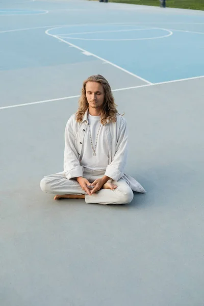 Полная длина стильный человек с длинными светлыми волосами медитируя в легкой позе йоги на открытом воздухе стадиона — стоковое фото