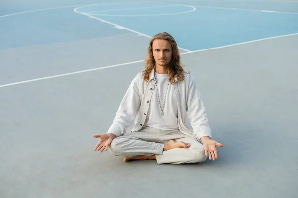 Полная длина длинноволосый мужчина в белой одежде медитирует в легкой позе, сидя на открытом стадионе — стоковое фото