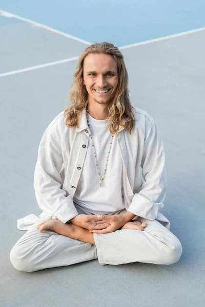Young and carefree man in stylish linen clothes sitting in lotus pose and smiling at camera on stadium — Stock Photo