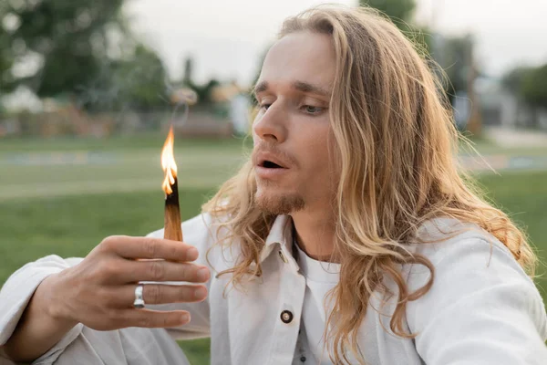 Homme aux cheveux longs en vêtements blancs soufflant à la combustion palo santo bâton — Photo de stock