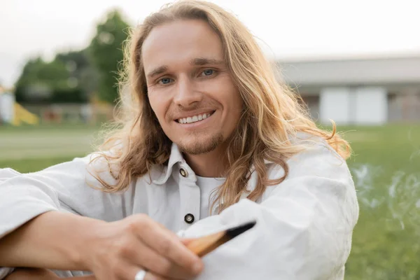 Беззаботный длинноволосый мужчина держит ароматный пало санто палкой и смотрит в камеру на улице — стоковое фото