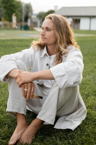 Calm barefoot man in white linen clothes looking away while sitting with aromatic palo santo stick outdoors — Stock Photo
