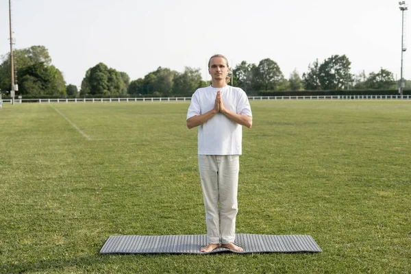 Homme pieds nus en pantalon de lin méditant en pose de montagne avec des mains priantes sur tapis de yoga sur pelouse verte — Photo de stock
