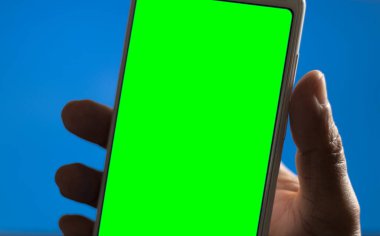 Male hand holding a white mobile phone or cellular phone on blue background. Natural light. Green screen.